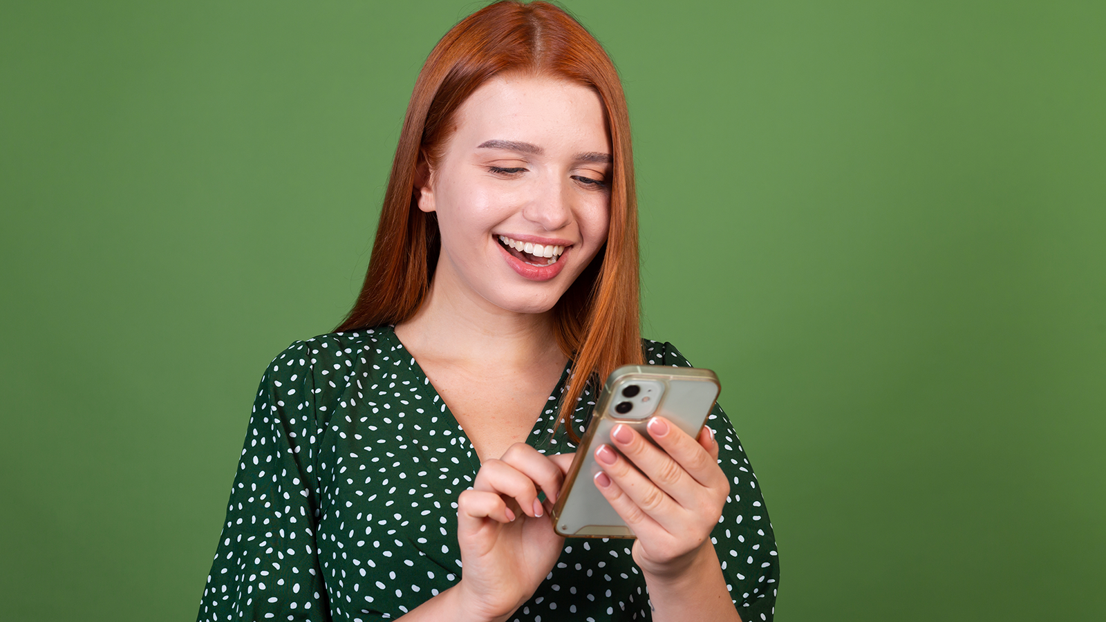 Uma mulher ruíva, branca, com uma blusa verde escura de bolinhas brancas, sorrindo e segurando um celular.