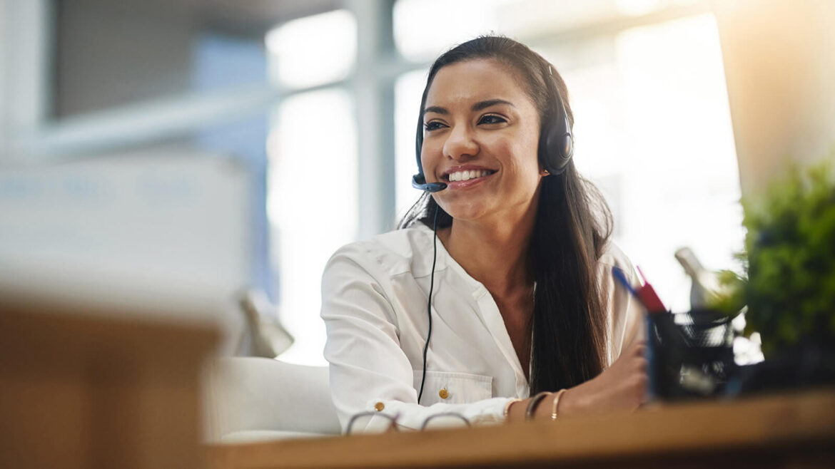 CRM no WhatsApp - Imagem de uma mulher negra de cabelo liso, cor castanha, vestida com uma blusa branca, sentada numa mesa, sorrindo, usando um headphone com o microfone, simbolizando trabalhar na área de atendimento ao cliente.