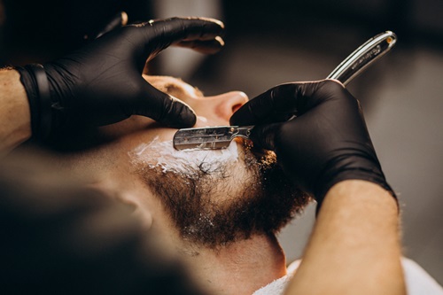  WhatsApp business para barbearia - Imagem de um barbeiro fazendo a barba de um homem branco, com camisa branca e pelos da barba escuros.