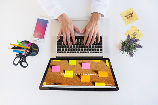 Marketing no WhatsApp - Imagem ilustrando o tema onde tem um notebook sobre uma mesa branca com post its colados na tela. Ao redor do computador, tem um celular, porta lápis e duas mãos como se estivessem digitando algo no notebook.