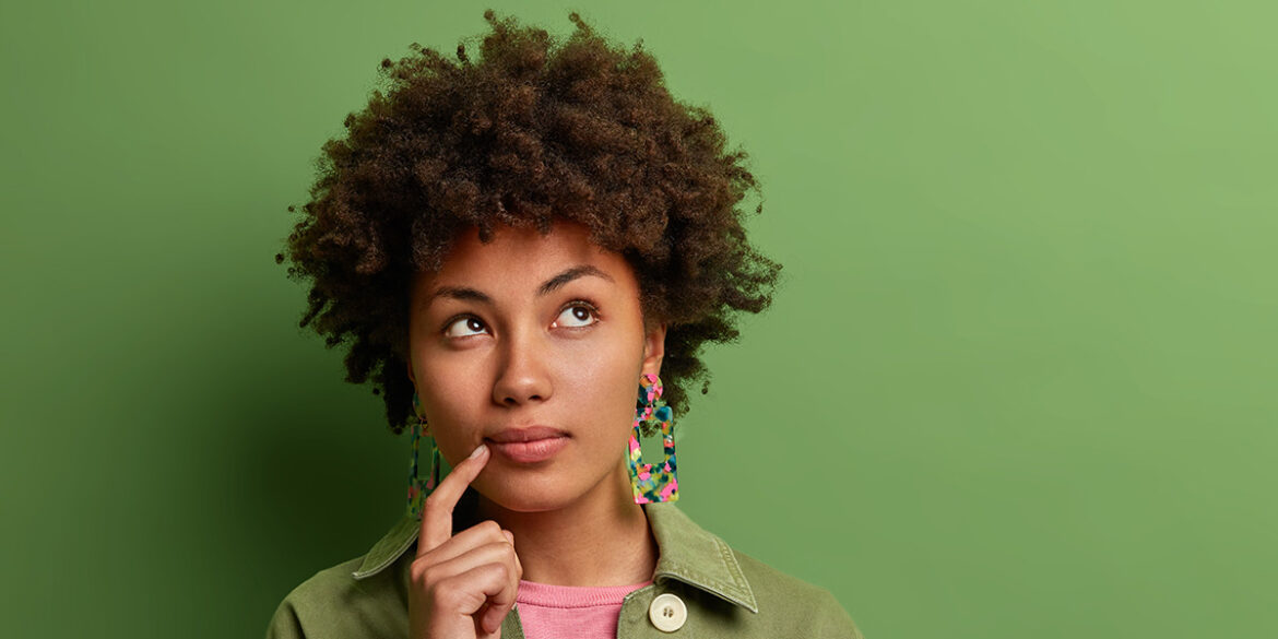 Alternativas para WhatsApp - A imagem contém um fundo verde musgo e, no centro, uma mulher negra de cabelo escuro, crespo, usando uma camisa de botão verde e, por dentro da camisa, uma blusa rosa. A mulher está com o dedo indicador no canto da boca, expressando estar pensativa.