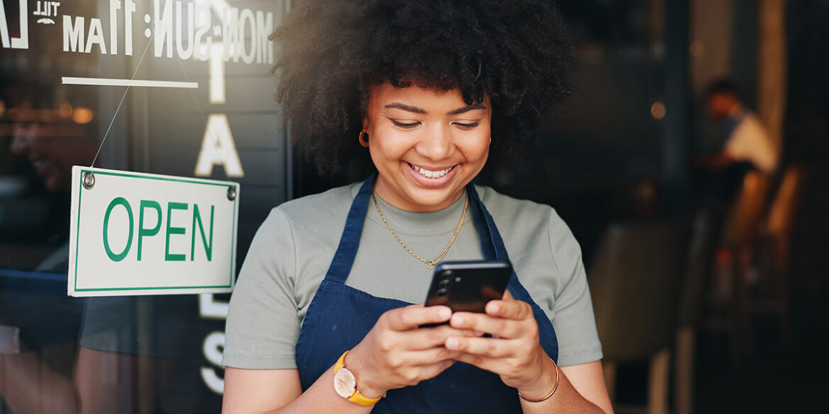 Branding no Whatsapp - Mulher negra de cabelo crespo, vestindo uma camisa cinza e avental azul escuro, segurando um celular preto e sorrindo enquanto olha pra tela.