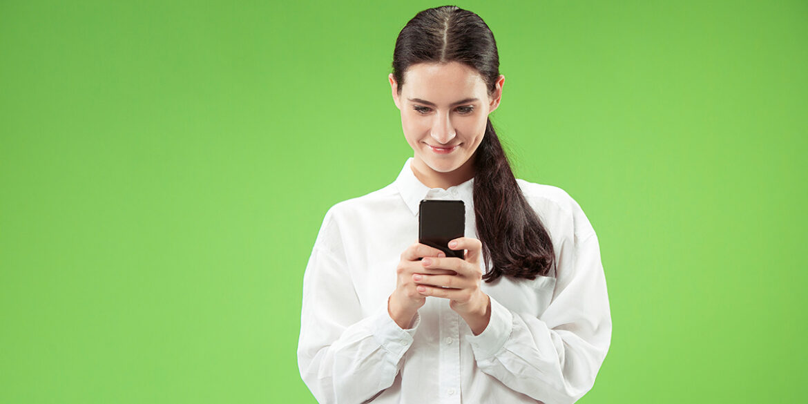 Campanhas para WhatsApp Marketing - A imagem que ilustra o tema tem o fundo verde e, no centro, uma mulher branca, de cabelo escuro liso, usando uma camisa de botão branca, segurando um celular.