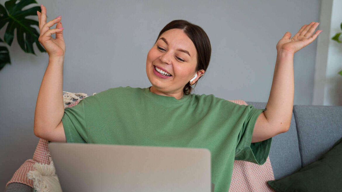 CRMs para o WhatsApp - Imagem ilustrando o tema descrito com uma mulher branca, de cabelo castanho, sentada no sofá que fica numa sala, de fundo, com um computador no colo, vestindo uma camisa verde, sorrindo e com as mãos levantadas na altura do ombro expressando alegria com algo que está vendo ou fazendo no computador.