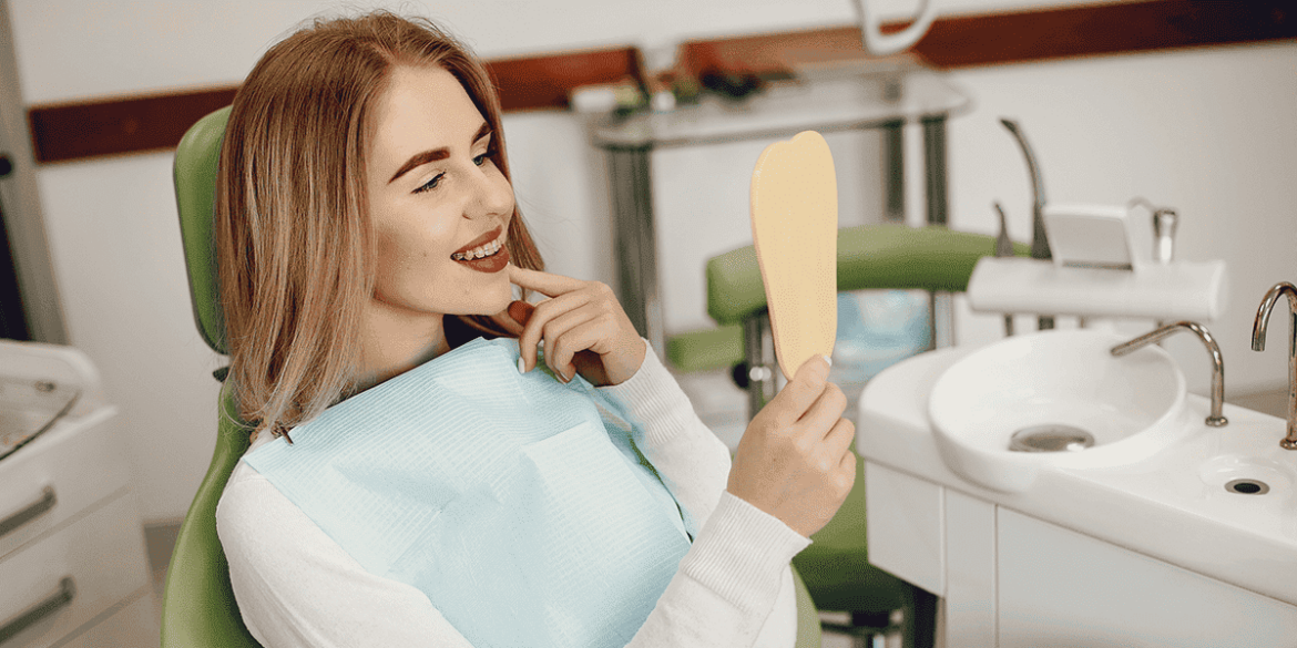 WhatsApp business para dentistas - uma mulher branca de cabelo liso, na cor castanho claro com mechas loiras, sentada numa cadeira de dentista, olhando para o espelho e sorrindo. De fundo, tem o consultório odontológico.