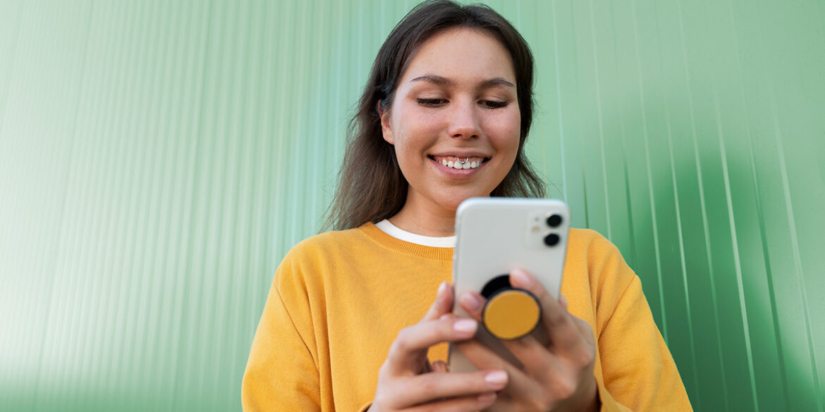 WhatsApp no omnichannel - Mulher parda de cabelo liso escuro, usando uma camisa amarela com gola branca, segurando um celular branco com as duas mãos e sorrindo olhando pra ele. Ela está sob um fundo verde.