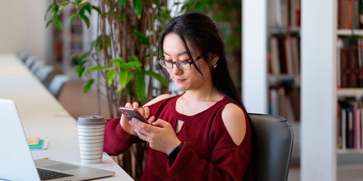 Mulher de blusa vermelha e oculos de grau, com o celular na mão e mexendo nele, um notebook aberto na mesa e um café na sua frente.