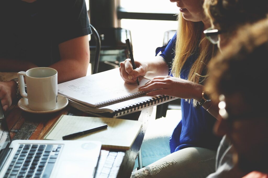 Automação de mensagens no meio empresarial - a imagem que ilustra o parágrafo tem um ambiente externo com uma mesa, onde tem pessoas sentadas em reunião. Na mesa tem um computador, uma xícara, bloco de anotação, caderno e canetas. Uma das pessoas que aparecem na foto, em destaque, é uma mulher branca, de cabelo longo loiro, usando uma camisa azul com calça jeans e escrevendo no caderno.