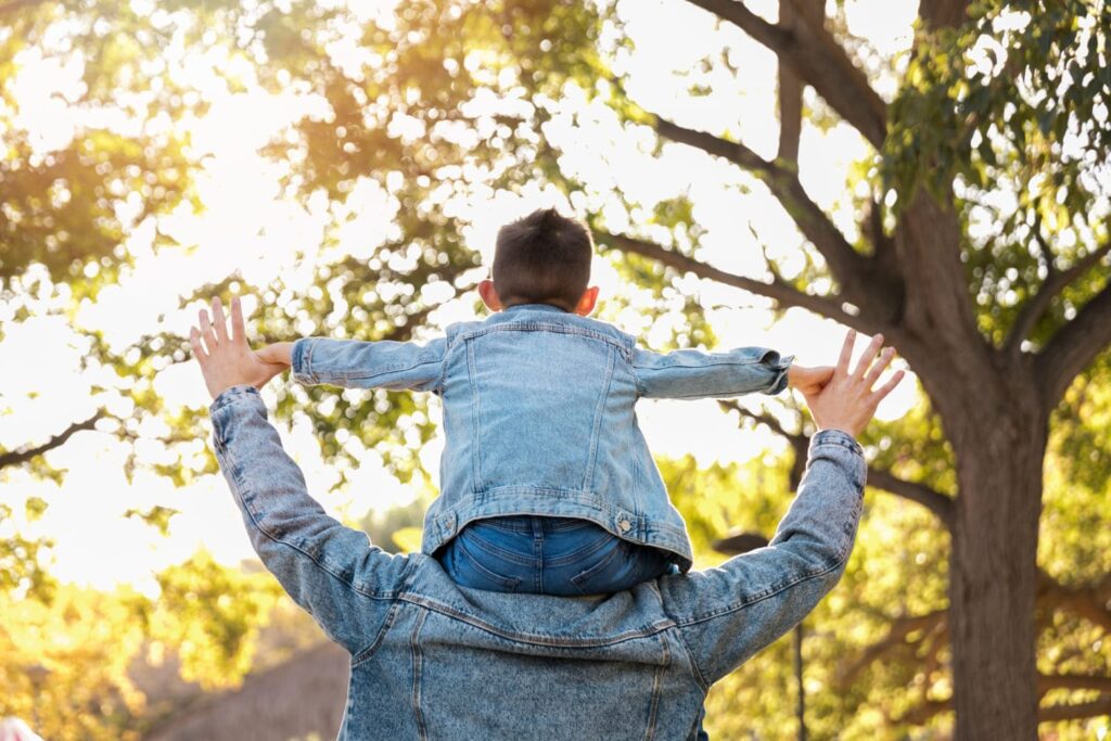 Campanha de dia dos pais - a imagem que ilustra o tópico tem um ambiente ao ar livre, arborizado e, no meio, tem um pai segurando o seu filho sobre os ombros.