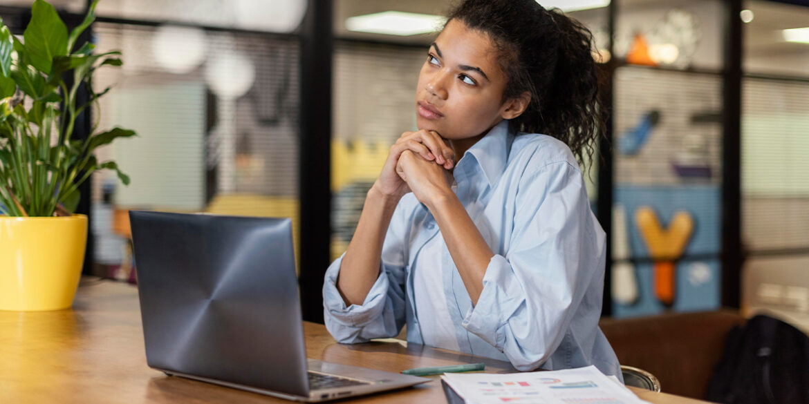 Atendimento no WhatsApp - a imagem que ilustra o tema tem o fundo de um escritório e, em destaque, tem uma mulher negra, de cabelo preto preso com um rabo de cavalo, usando uma blusa de botão azul, sentada em frente uma mesa com o notebook aberto e as mãos no rosto fazendo expressão de pensativa.