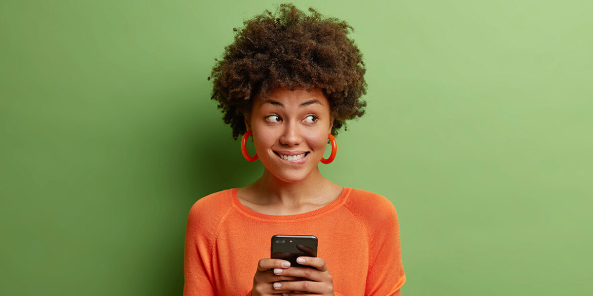 Campanhas no WhatsApp - A imagem que ilustra o tema tem o fundo verde com uma mulher, negra, de cabelo curto, cacheado, usando uma blusa laranja, segurando um celular com as duas mãos e olhando para o lado fazendo expressão de surpresa.