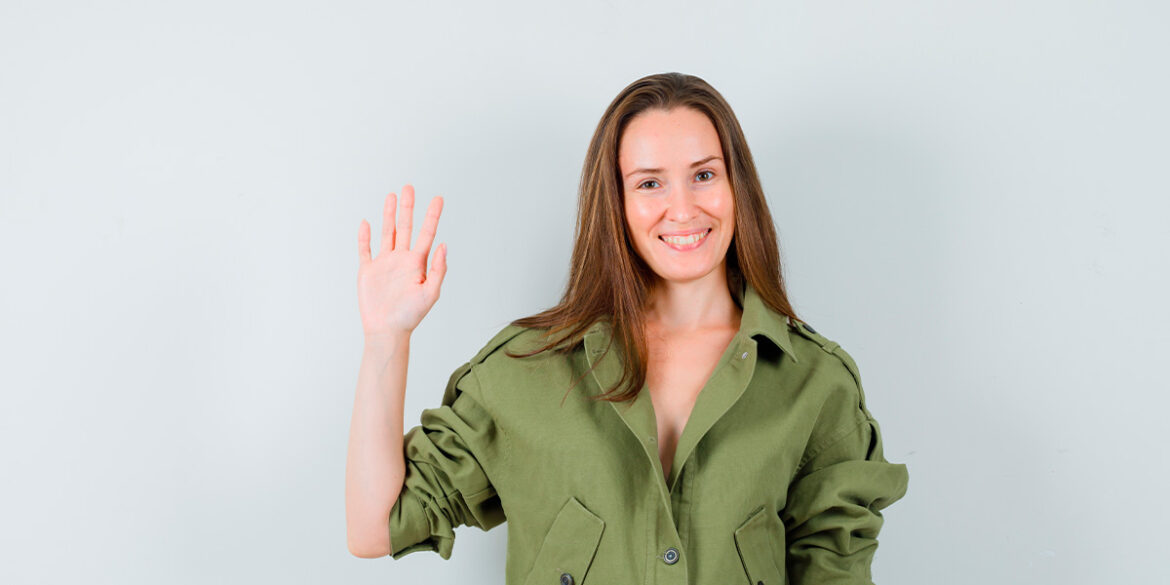 Mensagem de saudação no WhatsApp - A imagem que ilustra o tema tem o fundo branco com uma mulher, no meio, em destaque, posando para a foto, usando uma camisa de botão verde com uma das mãos levantadas acenando expressando saudação. A mulher é branca de cabelo liso castanho.