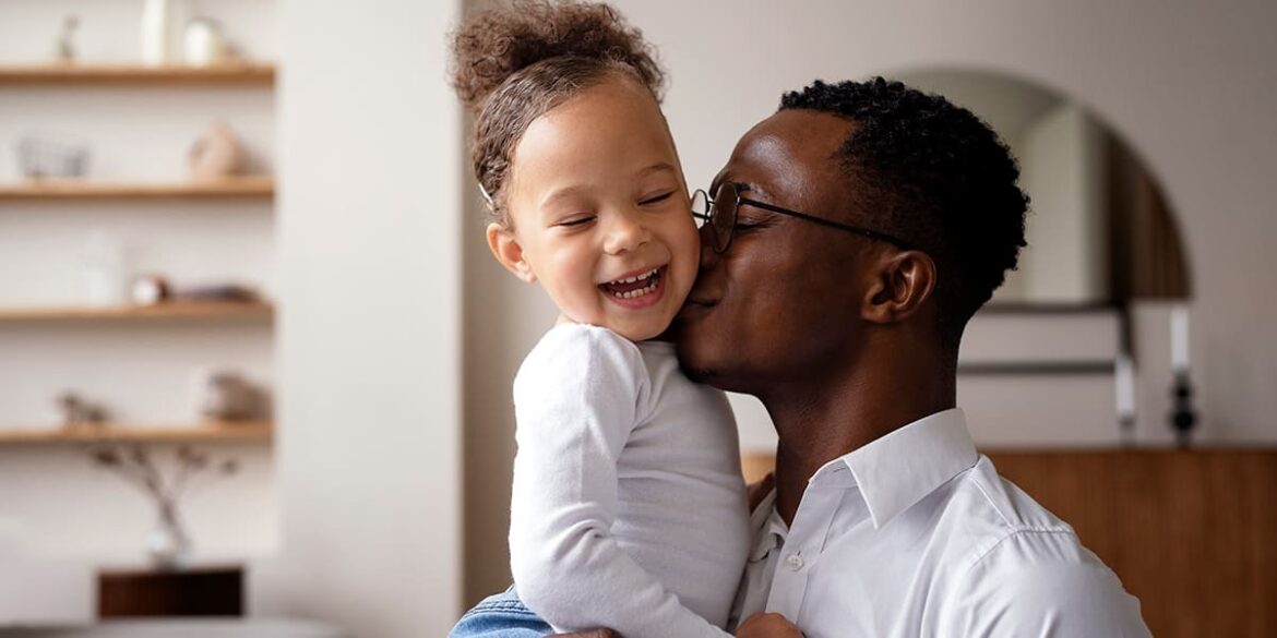 Campanha de dia dos pais - Pai segurando filha no colo e dando um beijo na sua bochecha e ela sorri.