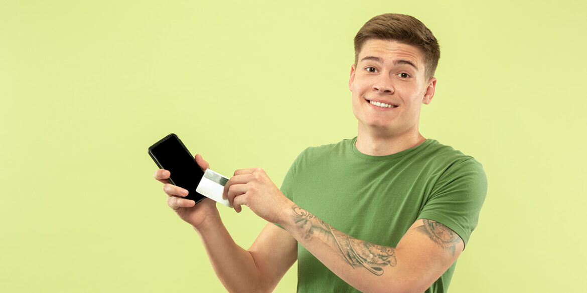 Pix no WhatsApp Business - a imagem que ilustra o tema tem o fundo verde, com um jovem rapaz, branco, de braços tatuados, usando uma camisa verde, aparece segurando um celular e um cartão, posando para a foto sorrindo.