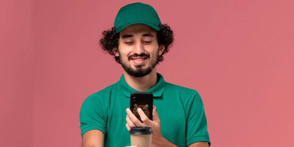 Respostas automáticas no WhatsApp - A imagem que ilustra o tema tem o fundo rosa claro e, no meio, em destaque, tem um homem, branco, de cabelo curto cacheado, vestindo uma camisa e boné verde, segurando um celular e sorrindo enquanto olha para o aparelho.