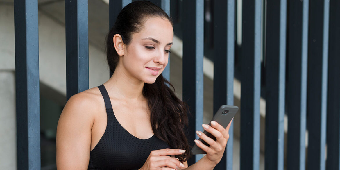 WhatsApp Business para Academias - a imagem que ilustra o tema tem o fundo de um estabelecimento e, em destaque, tem uma mulher, branca, de cabelo preso no rabo de cavalo, usando um top de treino preto, segurando uma garrafa numa mão e um celular na outra mão.