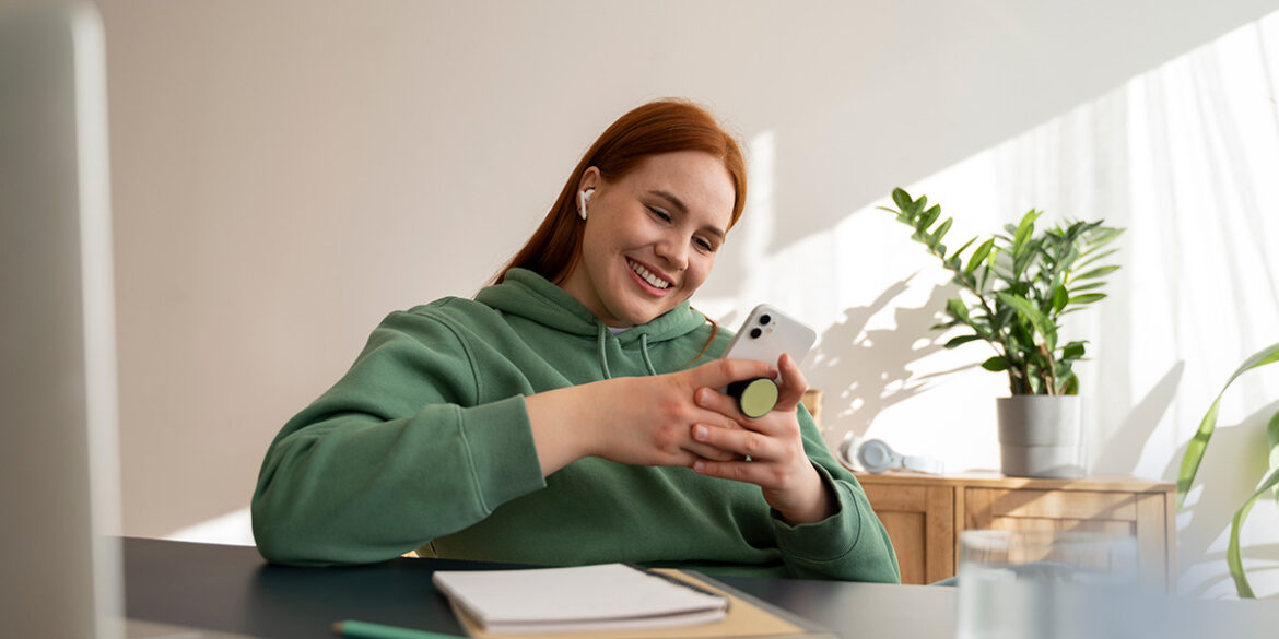 WhatsApp Business para freelancers - A imagem que ilustra o tema tem uma mulher branca, de cabelo ruivo liso, usando um moletom verde, sentada numa cadeira de madeira em frente a uma mesa, segurando celular e sorrindo. Na mesa, tem um caderno e um livro, apoiados, e ao lado uma caneta.