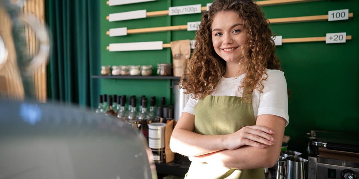 WhatsApp business para restaurantes - a imagem que ilustra o tema tem uma mulher branca, de cabeços ondulados castanhos, usando uma blusa branca e avental verde, atrás de um balcão de pagamento de um estabelecimento. Atrás dela, tem uma parede com bebidas e outros itens a venda.