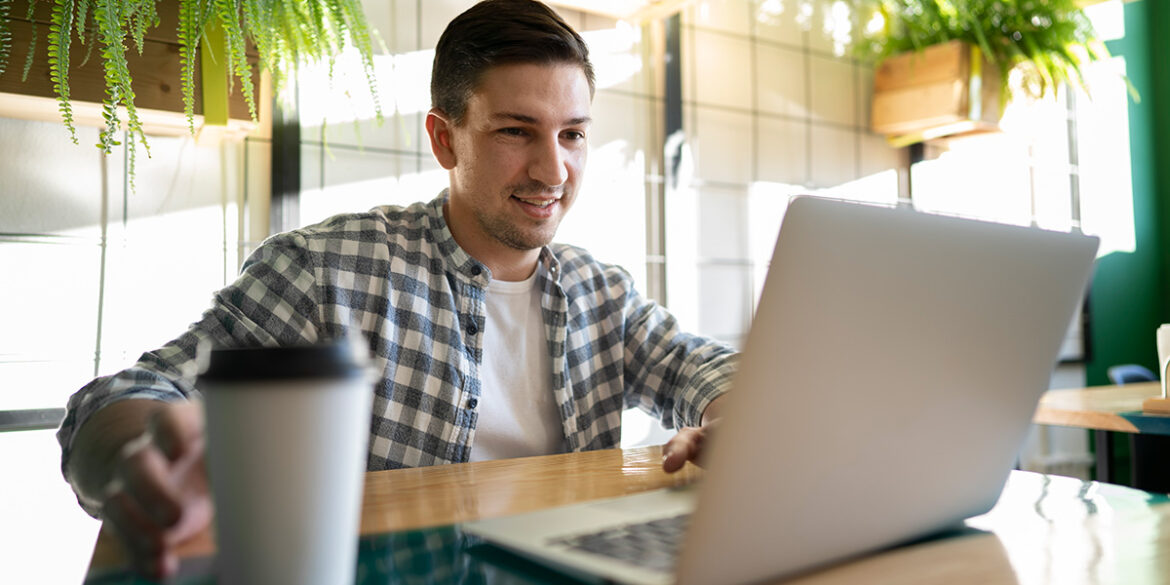 WhatsApp para o seu time - a imagem que ilustra o tema tem o fundo de uma sala com uma mesa de madeira, onde um homem branco, está sentado, em frente a um computador com uma camisa de botão xadrez por cima de uma camisa branca