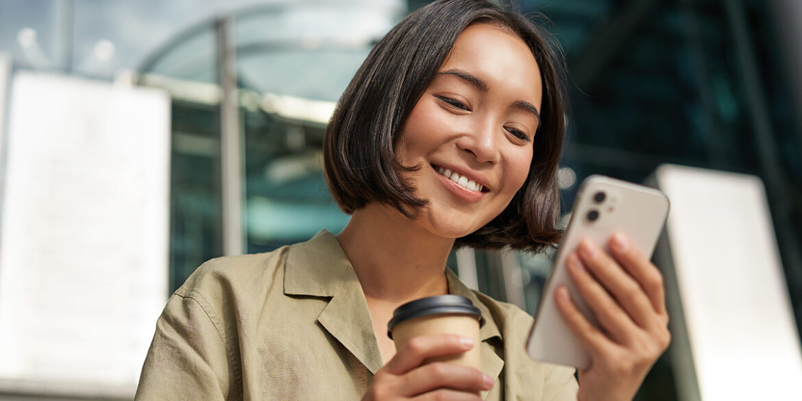 fechamento de vendas no WhatsApp - a imagem que ilustra o tema tem uma mulher parda, de cabelo curto, liso e preto com traços asiáticos segurando um celular e um copo de café. Ela está sorrindo enquanto olha para o celular e de fundo aparece um estabelecimento público.