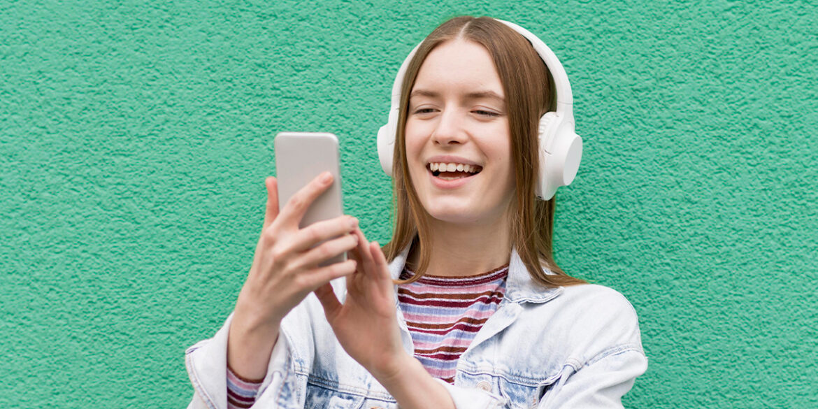 Como criar conteúdos em áudio no WhatsApp - A imagem que ilustra o tema tem o fundo verde e, no meio, tem um modelo branca de cabelo liso castanho, usando uma jaqueta jeans por cima de uma blusa listrada, usando um headphone, segurando um celular e sorrindo para ele.