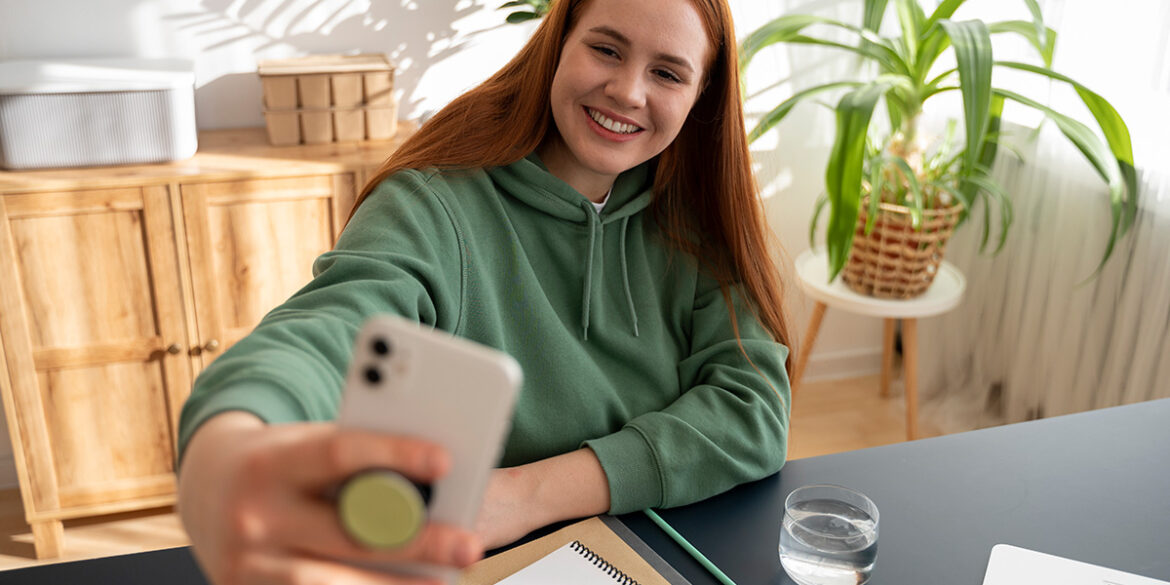 Como criar conteúdos em vídeo - A imagem que ilustra o tema tem o fundo de uma sala com uma mulher sentada em frente a uma mesa, segurando um celular como se estivesse fazendo a captura de um vídeo ou imagem dela mesma.