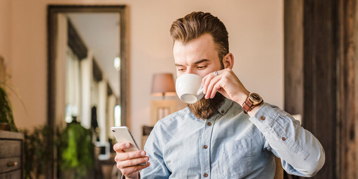 Mensagem de saudação para barbearias - A imagem que ilustra o tema tem o fundo de uma sala onde tem um homem, branco, sentado, tomando café enquanto mexe no celular.