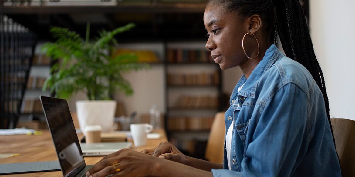 Upselling e Cross-selling - a imagem que ilustra o tema tem o fundo de uma sala de escritório e, em foco, tem uma mulher negra, usando uma jaqueta jeans por cima de blusa branca, sentada em frente um computador.