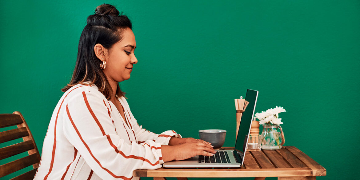 Guia de atendimento no WhatsApp - a imagem que ilustra o tema tem o fundo verde escuro e aparece uma mulher sentada num banco de madeira em frente uma mesa, mexendo no computador digitando e sorrindo.