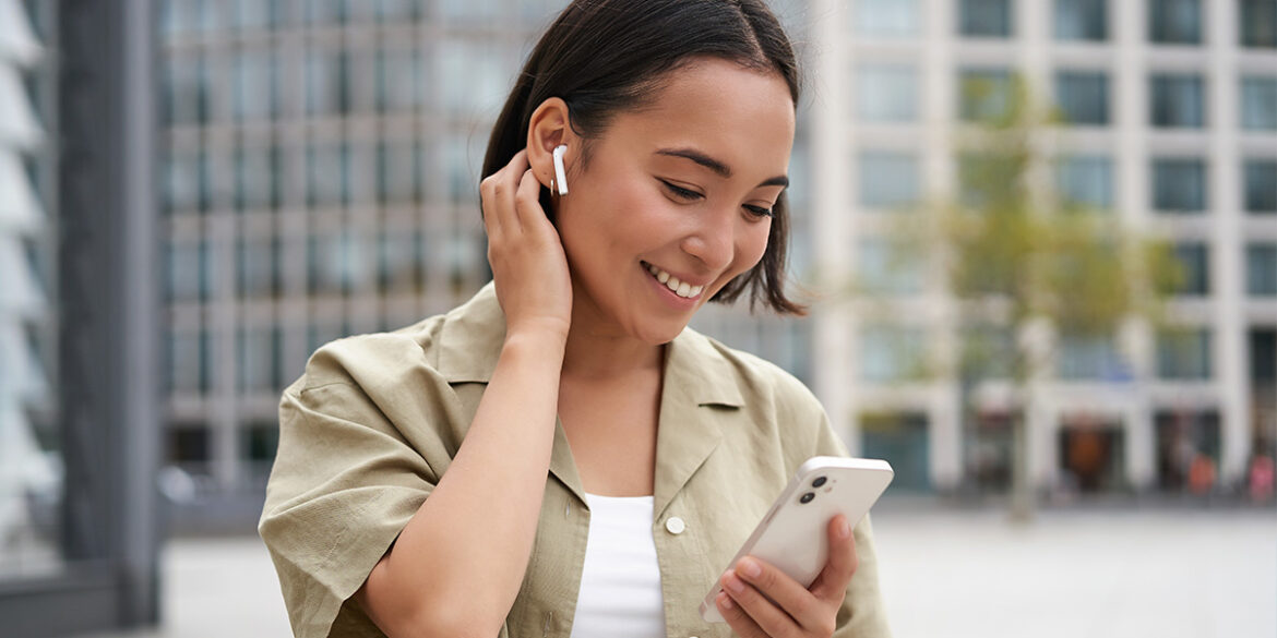 Marketing de Voz - a imagem que ilustra o tema tem o fundo de uma rua e, em destaque, te uma mulher, negra, de cabelo curto liso, usando uma camisa de botão bege sobre uma blusa branca, segurando um celular e sorrindo.