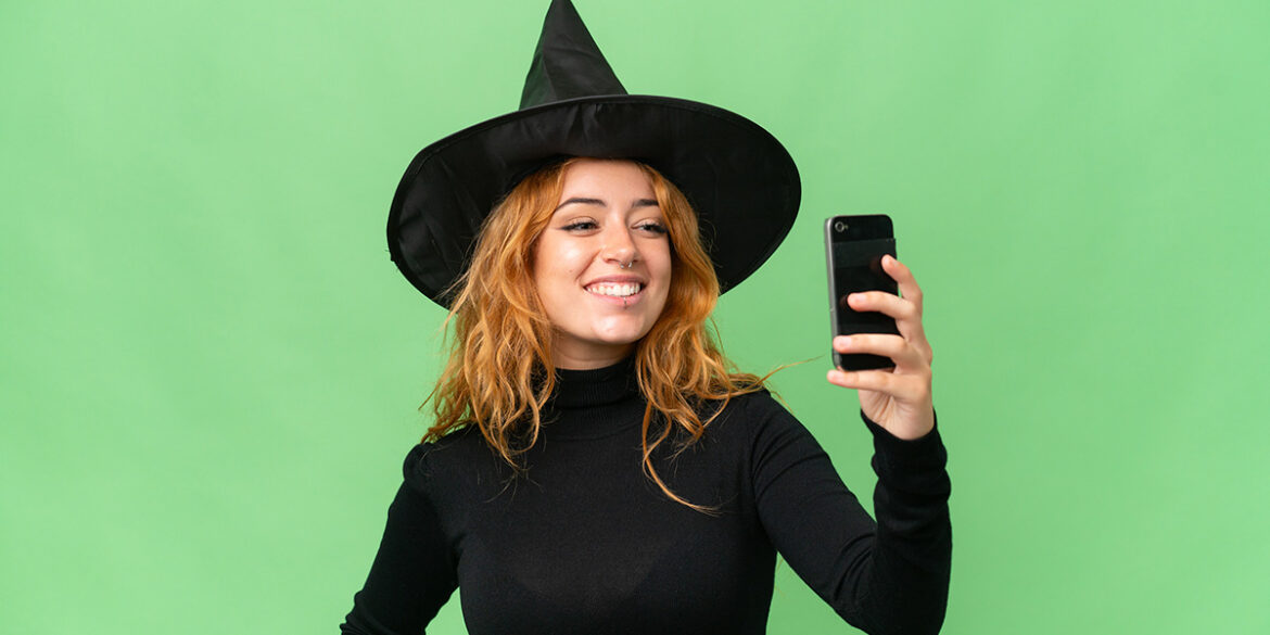 Campanha de Halloween no WhatsApp - A imagem que ilustra o tema tem o fundo verde e, no meio, uma mulher branca, de cabelo ruivo, fantasiada de bruxa, segurando um celular e sorrindo como se tivesse tirando uma foto dela mesma.