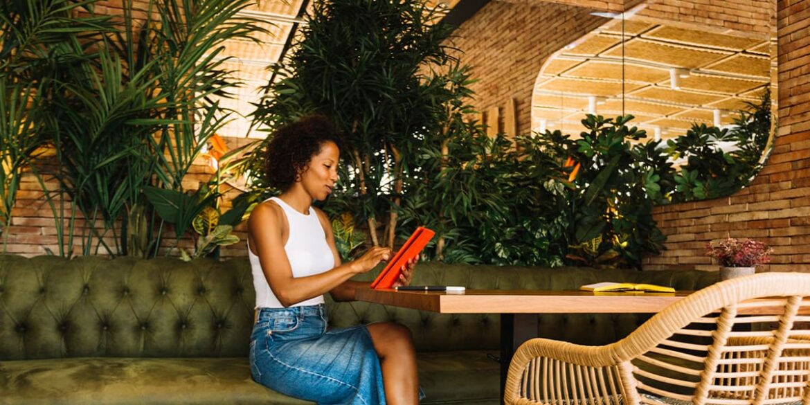 Etiquetas fundamentais para restaurantes - A imagem tem o fundo de um estabelecimento público com muitas plantas e um espaço com mesa e cadeiras onde tem uma mulher, negra, de cabelo cacheado, usando uma blusa branca e saia jeans, sentada numa das cadeiras em frente a mesa, segurando um tablet e mexendo nele.