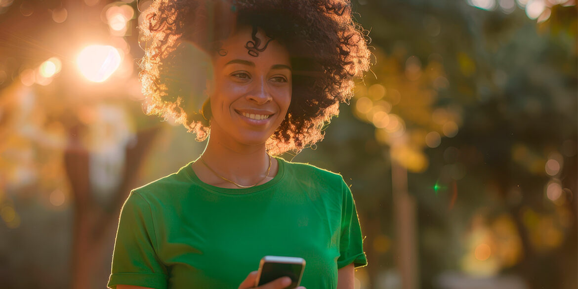 Marketing Conversacional no WhatsApp - A imagem que ilustra o tem tem o fundo de um ambiente ao ar livre, arborizado, com uma mulher negra, de cabelos cacheados, usando uma blusa verde, aparece sorrindo e segurando um celular.