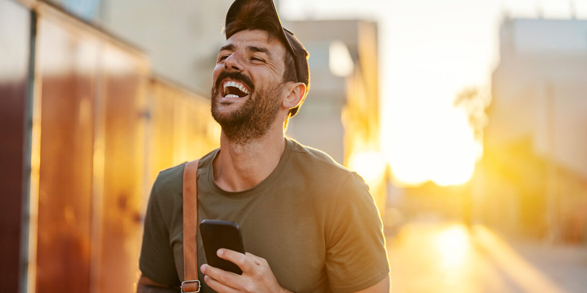 Memes no WhatsApp Business - A imagem que ilustra o tema tem o fundo de uma rua e, em destaque, um homem usando uma camisa de manga longa, de chapéu, segurando um celular e sorrindo.