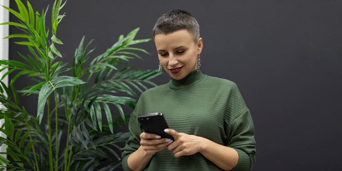 WhatsApp Web - A imagem que ilustra o tema tem o fundo escuro com uma planta na cor verde e uma mulher, de cabelo curto, usando uma camisa de manga longa verde, olhando e segurando um celular.