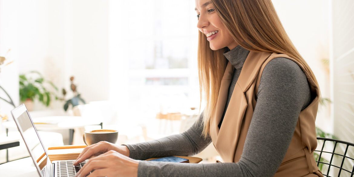 Marketing de Conteúdo no WhatsApp - A imagem que ilustra o tema tem uma mulher sentada numa mesa, mexendo num computador e sorrindo. Ao lado tem uma xícara de café e, de fundo, tem aparentemente um ambiente de cafeteria.