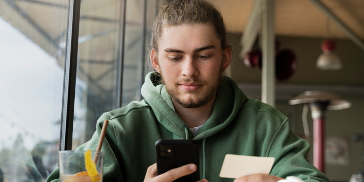 Homem numa lanchonete, com o celular numa mão e o cartão na outra, finalizando uma compra.