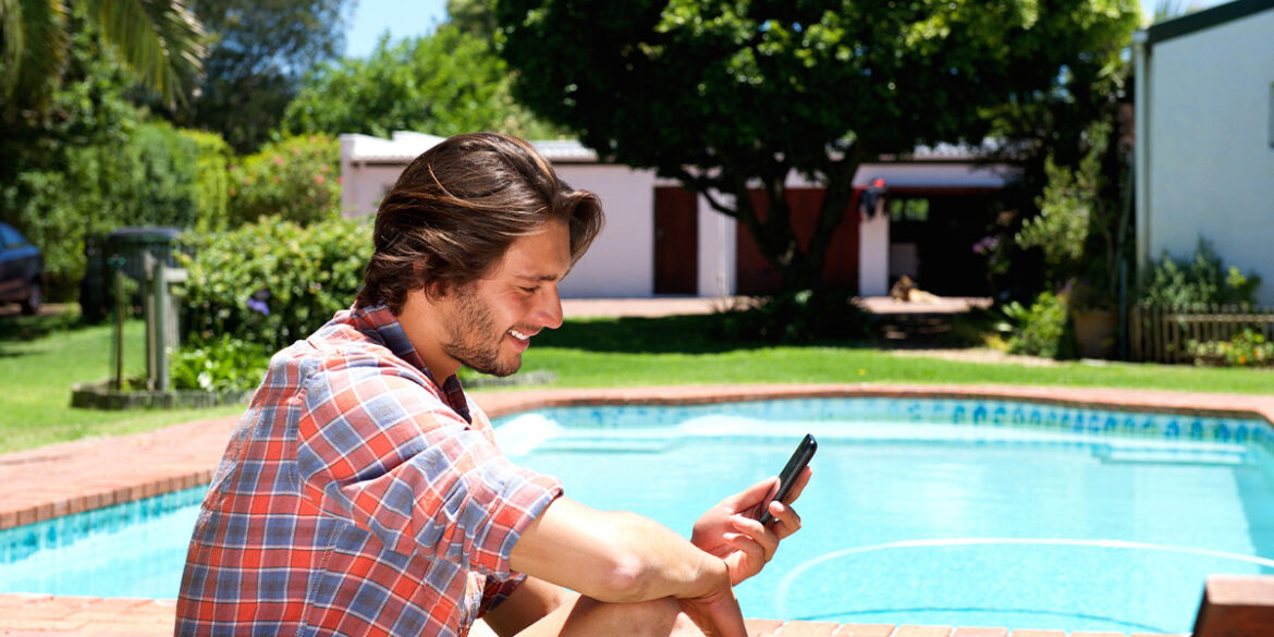 Homem na piscina do condomínio, mexendo no celular e sorrindo.