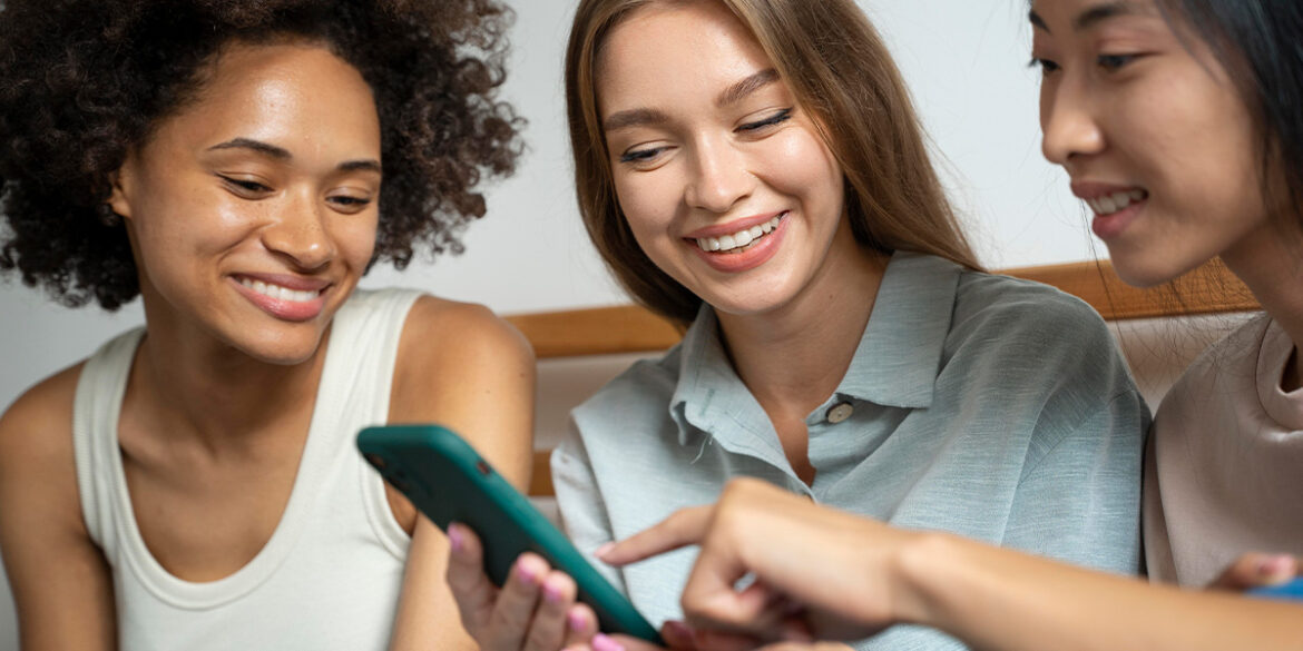 Três meninas mexendo no celular, conversando e sorrindo.